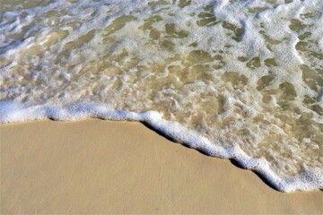 A wave rolls back from the shore, exposing light, clean sand and foaming at its edge, viewed from above