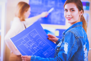 Two young woman standing near desk with instruments, plan and laptop.