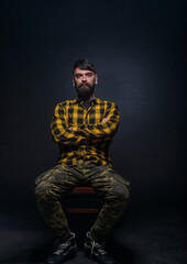 Tattooed man with a beard wearing checkered shirt is posing seated on a chair in a studio