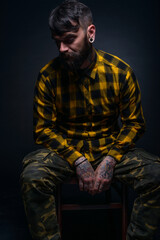 Fototapeta premium Tattooed man with a beard wearing checkered shirt is posing seated on a chair in a studio