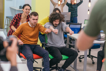 Obraz premium Team building and office fun.Four young cheerful businesspeople in smart casual wear having fun while racing on office chairs and smiling..