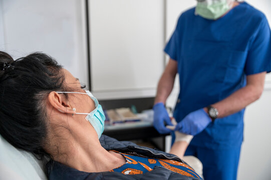 Woman Covered With Surgical Protection Mask Is Treated By A Doctor In An Outpatient Clinic During The Coronavirus Period. Bandaging A Hand In The Emergency Room Of A Hospital After A Home Injury.