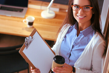 Young woman standing near desk with laptop holding folder and cup of coffee