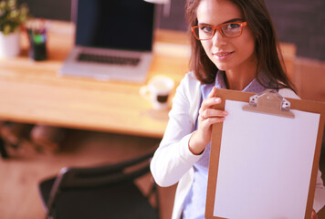 Young woman standing near desk with laptop holding folder and cup of coffee. Workplace. Business...