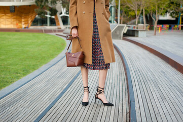 Young fashionable woman wearing beige wool coat, pleated midi skirt and black high heel shoes. She is holding leather handbag in hand. Street style. 