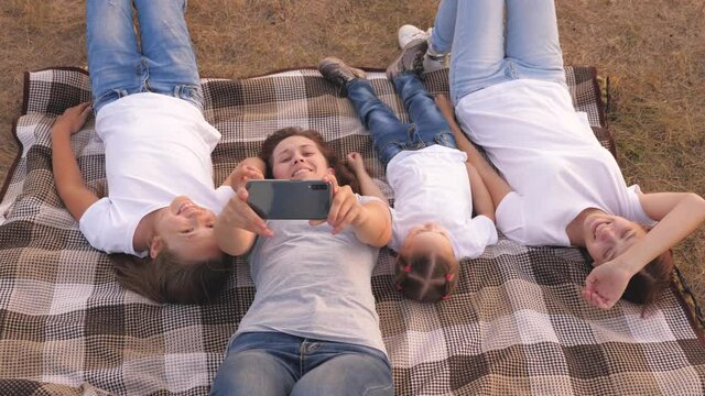 Mom and happy children are photographed on a smartphone in park. Mother and daughters take a selfie while traveling, lying on bedspread. happy family and smartphone. funny kids and parents on vacation