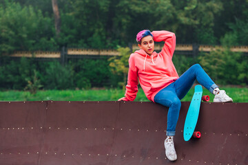 Portrait of a trendy pretty young gir with short colored hair sitting next to the skateboard court with her plastic skateboard.