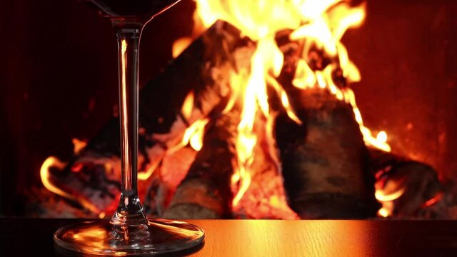 Glass of red wine and birch logs of firewood in burning fire in fireplace, close-up