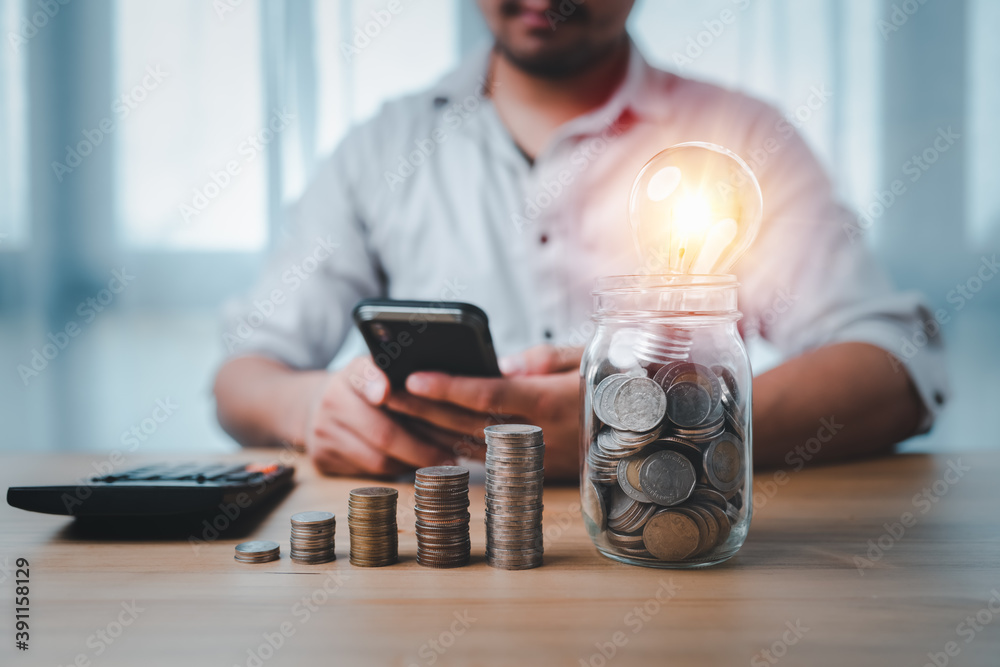 Leinwandbilder coins stacking on desk and lightbulb in jar with businessman use smartphone and calculator. saving e