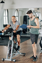 Fototapeta na wymiar Concentrated mature man riding on stationary bike when trainer writing results in document