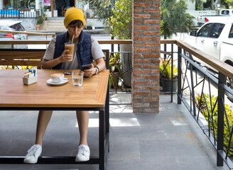 Young woman in yellow hat drinking ice coffee and typing message on phone at cafe.