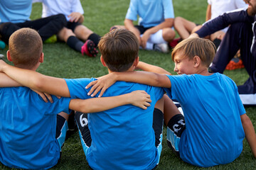 young kids football team embrace each other on football field before match, support, have talk, sit on grass together