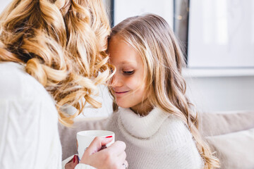 Mother and daughter in light sweaters are sitting on the sofa, the room is decorated for the new year and Christmas, hugs, conversations, smiles, kisses, waiting for a gift and magic