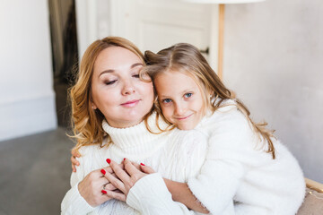 Mother and daughter in light sweaters are sitting on the sofa, the room is decorated for the new year and Christmas, hugs, conversations, smiles, kisses, waiting for a gift and magic