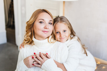 Mother and daughter in light sweaters are sitting on the sofa, the room is decorated for the new year and Christmas, hugs, conversations, smiles, kisses, waiting for a gift and magic