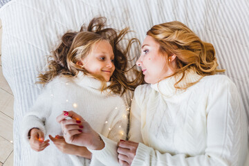 Mom and daughter blonde hair and white sweaters lie on the bed, light up, say toys for Christmas and new year tree, Christmas tree decoration