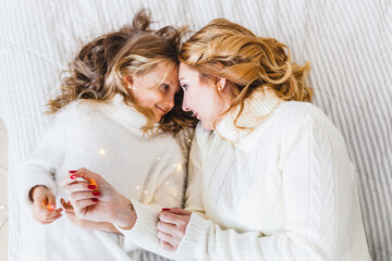 Mom and daughter blonde hair and white sweaters lie on the bed, light up, say toys for Christmas and new year tree, Christmas tree decoration