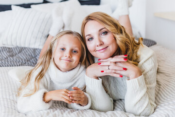 Mom and daughter blonde hair and white sweaters lie on the bed, light up, say toys for Christmas and new year tree, Christmas tree decoration