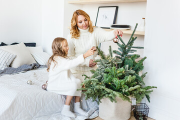 Mom and daughter blonde hair and white sweaters lie on the bed, light up, say toys for Christmas and new year tree, Christmas tree decoration