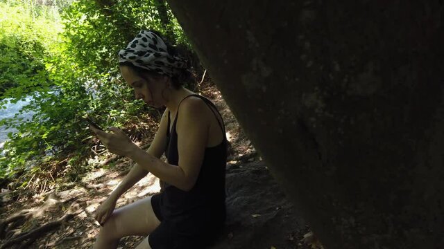 Young woman sitting on a stone by the Banias river, concentrated at her phone screen.