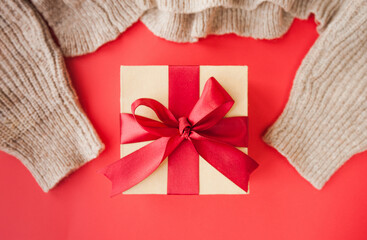 beige box with red festive satin ribbon and beige knitted sweater on gradient red background. minimalism greeting card for holiday, selective focus