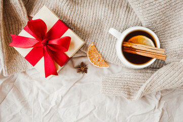 Fototapeta na wymiar cozy Christmas hygge style card. porcelain tea cup with orange slice, cinnamon sticks, anise star on beige crumpled blanket, gift with red ribbon and sparkling garland on background, selective focus