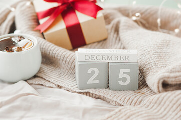 wooden square calendar cubes with the date December 25 on beige cozy crumpled blanket, gift with red ribbon and sparkling garland on background, selective focus