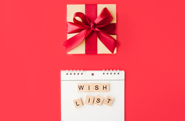 white spiral notebook and square lettering wishlist on red background, gift box made of Kraft paper with red satin bow,selective focus