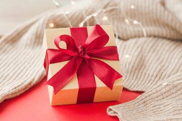 stylish square beige box with red festive satin ribbon on beige crumpled blanket and sparkling garland on red background, selective focus