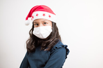 Fototapeta na wymiar Little girl wearing santa hat and face mask looking to the camera sad on white background.