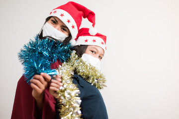Mother and daughter looking to the camera wearing santa hat and surgical mask holding tinsel sparkled on white background with copy space for text.