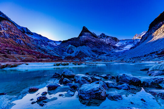 A river flowing through a valley in late autumn with snow capped mountains in the background, rocks in the foreground and the sun starting to rise