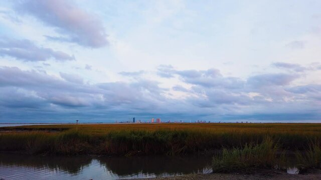 4K TIMELAPSE Atlantic City skyline in distance over waterway with mostly cloudy sky day to night with clouds turning pink, purple and blue with blur