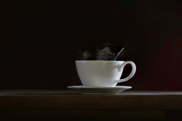 Coffee in white cup put on saucer and on the plank which has dim light and dark wall