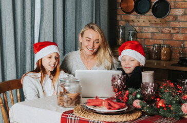 Family mother and two children in Santa hats in the kitchen decorated for Christmas having video chat with relatives at served table
