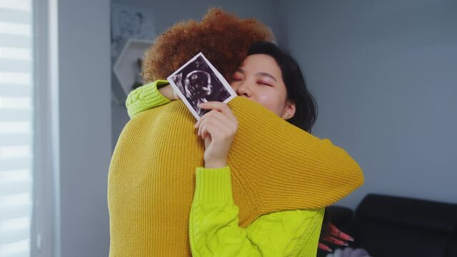 Multiracial lesbian couple expecting baby. Embracing with ultrasound baby picture. Maternity support. High quality 4k footage