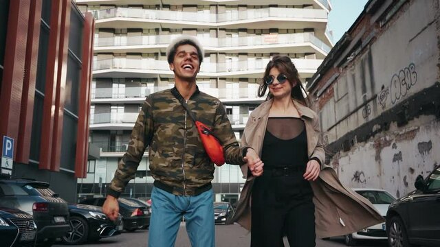 Front view of multicultural couple in trendy clothing holding hands and running at modern city center. Concept of relationship, fun and love.