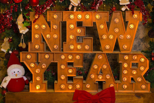 "New Year" inscription made of wood with glowing lanterns and a toy snowman next to it: festive atmosphere, soft focus, Christmas background in the frame of the decorated branches of a Christmas tree