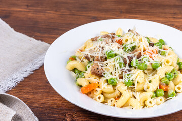 Traditional italian chifferini pasta with peas, carrots and toasted pancetta, sprinkled with grated cheese on a plate on a rustic wooden table, background