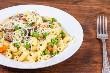 Traditional italian chifferini pasta with peas, carrots and toasted pancetta, sprinkled with grated cheese on a plate on a rustic wooden table, background