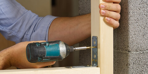 Craftman holds a portable screwdriver for fixing metal corners with screwing screws into the wood board.