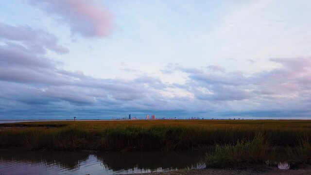 4K TIMELAPSE Atlantic City skyline in distance over waterway with mostly cloudy sky day to night with clouds turning pink, purple and blue