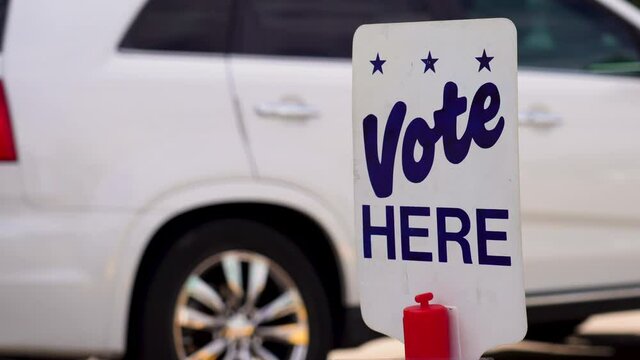 Blue Vote Here Sign Close up with People Driving Cars Walking in the Background