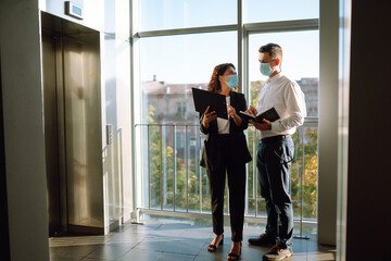 Two coworkers in protective face masks discussing project, sharing ideas, having pleasant conversation near elevator. Work in office during quarantine. Covid-19.