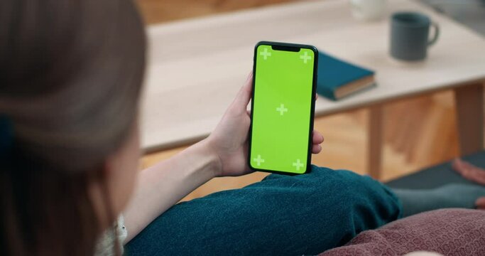 Backside view of woman showing i love you sign while looking at smartphone mock up screen. Female person having video call while sitting on couch at home. Concept of croma key.