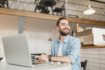 Smiling funny young man sitting alone at table in coffee shop cafe working or studying on laptop pc computer, relaxing in restaurant during free time indoors. Freelance mobile office business concept.