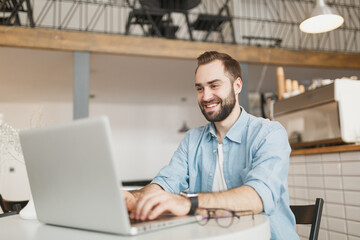 Smiling handsome young man sitting alone at table in coffee shop cafe working studying on laptop pc...