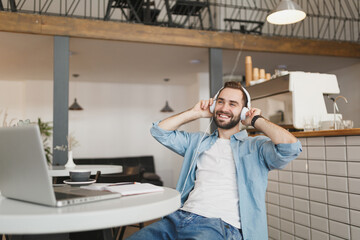 Smiling young man sitting alone at table in coffee shop cafe restaurant indoors working or studying on laptop pc computer listening music with headphones. Freelance mobile office business concept.