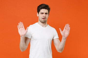 Displeased shocked worried nervous young man 20s in basic casual white t-shirt standing rising hands up showing palms looking camera isolated on bright orange colour wall background, studio portrait.