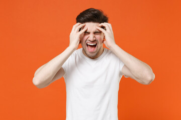 Fototapeta na wymiar Crazy frustrated young man 20s wearing basic casual blank white t-shirt standing put hands on head keeping eyes closed screaming isolated on bright orange colour wall background, studio portrait.